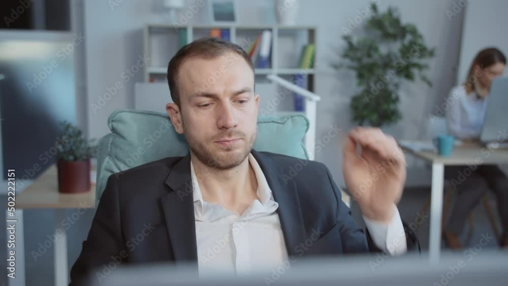 Portrait of young Caucasian businessman in black suit with hands behind head sitting in office leaning back in chair. Footage of handsome man sitting at computer, looking at monitor. Indoors Portrait of young Caucasian businessman in black suit with hands behind head sitting in office leaning back in chair. Footage of handsome man sitting at computer, looking at monitor. Indoors