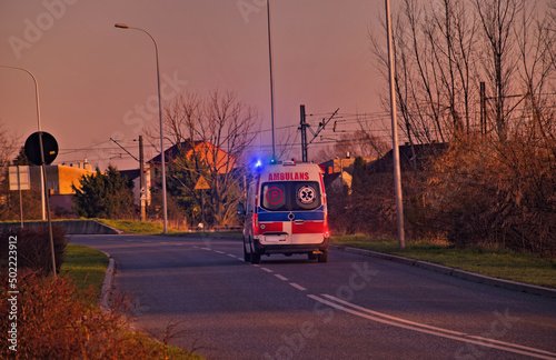 Ambulans na sygnale ( włączone migające lampy " koguty " ) jadący miejską ulicą , przy zachodzącym słońcu .