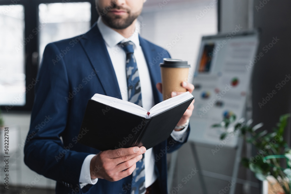 cropped view of blurred businessman with notebook and paper cup in office.