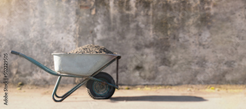 construction wheelbarrow with a pile of sand. a dirty construction wheelbarrow stands against a gray wall in the sun