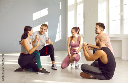 Fototapeta Naklejka Na Ścianę i Meble -  Young people having a chat after a fitness workout. Group of happy friends in sportswear sitting on the gym floor, talking, sharing their achievements, discussing news and smiling