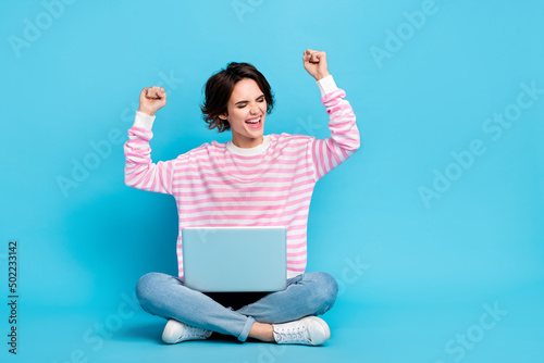 Photo of ecstatic pretty girl raise fists in victory achievement sit floor with laptop isolated on blue color background © deagreez