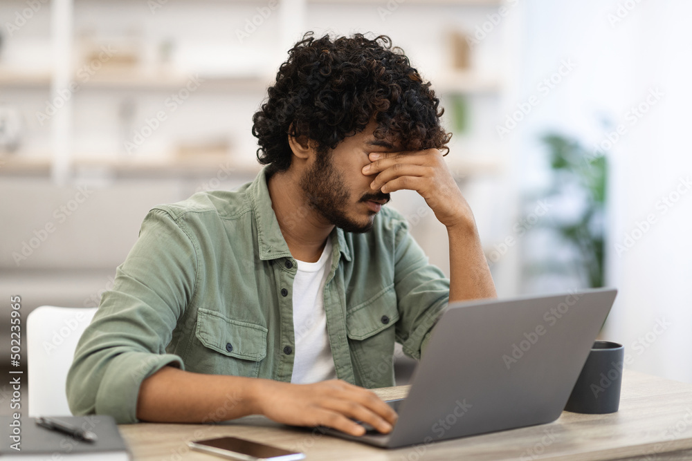 Tired dark-skinned guy sitting at workdesk in front of laptop Tired dark-skinned guy sitting at workdesk in front of laptop
