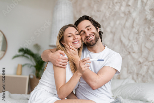 Happy young couple celebrating positive result of pregnancy test, hugging and cuddling on bed at home
