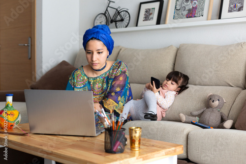 Young Muslim woman working at home with her laptop while her young daughter plays with a mobile phone. Single parent family.