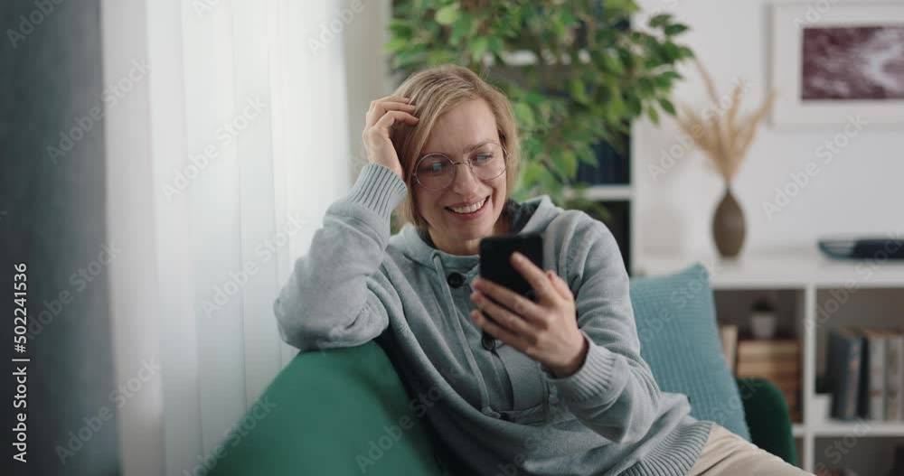 Happy mature woman in casual clothes sitting on sofa and scrolling news on modern cell phone. Concept of modern gadgets, people, and leisure at home. Happy mature woman in casual clothes sitting on sofa and scrolling news on modern cell phone. Concept of modern gadgets, people, and leisure at home.