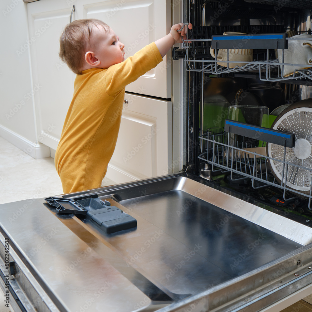 Baby boy toddler is standing by an open dishwasher with dirty dishes inside. Child safety issues in the home room, little kid Baby boy toddler is standing by an open dishwasher with dirty dishes inside. Child safety issues in the home room, little kid