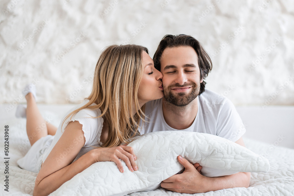Affectionate young married couple relaxing, hugging pillow, kissing on bed at home, feeling carefree in morning Affectionate young married couple relaxing, hugging pillow, kissing on bed at home, feeling carefree in morning