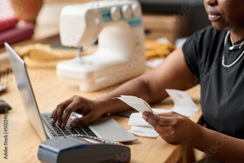 Hands of young African American woman with receit checking payment information and entering data while sitting in front of laptop photo