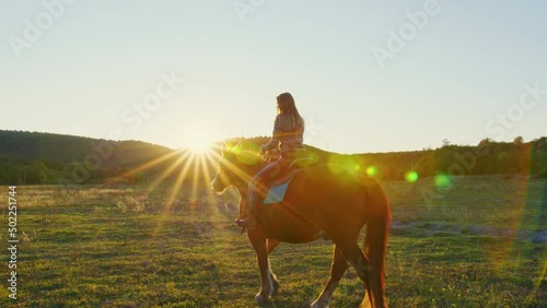 Beautiful girl rides horse in pasture surrounded by forests, hills. Young woman goes on horseback in countryside. Light and shadow. Sun shining above mountain. Horsewoman is on saddle. Palfrey walks photo