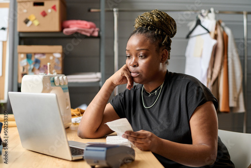 Young serious black woman with receit looking at laptop screen by workplace while checking payment of online clients photo
