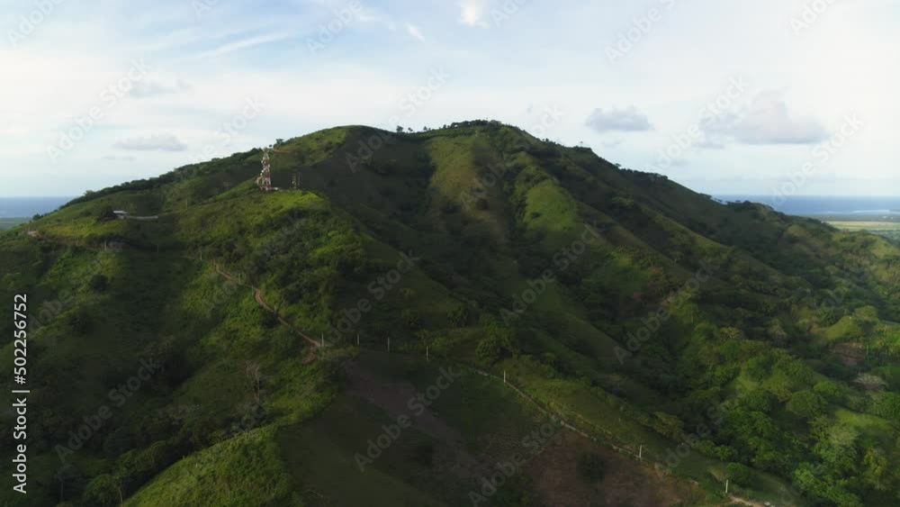 A picturesque mountain peak with a road against the background of the sea. Green slopes of the mountain in the rays of the morning sun. Landscape of wild nature. Blue cloudy sky over tropical coast. A picturesque mountain peak with a road against the background of the sea. Green slopes of the mountain in the rays of the morning sun. Landscape of wild nature. Blue cloudy sky over tropical coast.
