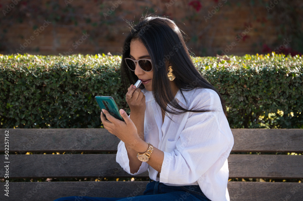 a beautiful young south american woman on holiday in europe, is sitting on a park bench painting her lips while using her mobile phone as a mirror. a beautiful young south american woman on holiday in europe, is sitting on a park bench painting her lips while using her mobile phone as a mirror.