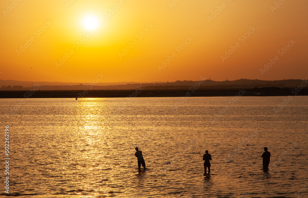Long shot of a sunset at the beach with silhouette of three fishermans