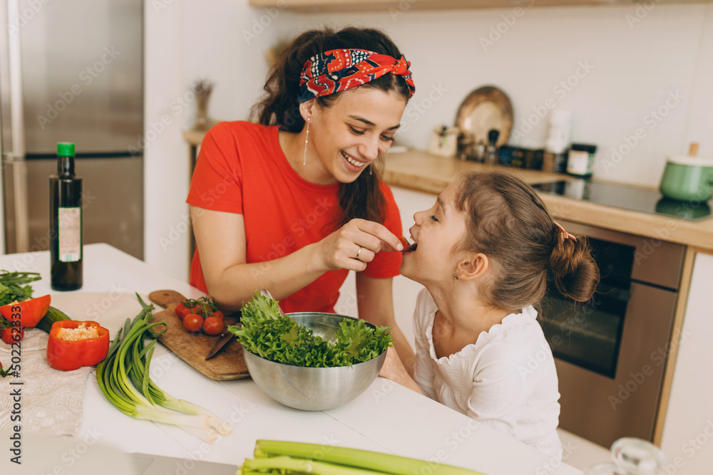 Happy mom and daughter at kitchen, cooking together, mother giving child cherry tomato to try, woman wearing red t-shirt and stylish bandana, girl dressed in white, vegetables and utensils on table Happy mom and daughter at kitchen, cooking together, mother giving child cherry tomato to try, woman wearing red t-shirt and stylish bandana, girl dressed in white, vegetables and utensils on table