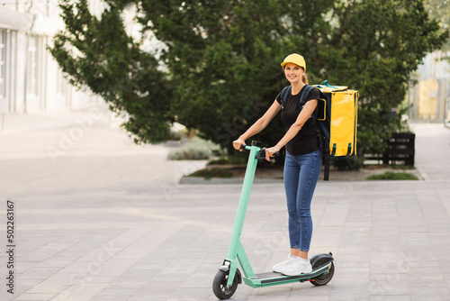 Beautiful young girl with a yellow bag on her back and cap riding an electric scooter down the street early in the morning. Online delivery service concept. Ecological, green transport