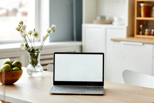 Minimal background image of white laptop screen on wooden table in kitchen decorated with fresh flowers and fruits, copy space
