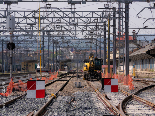 Train tracks under construction with red and white signs preventing traffic and a safety pole connecting the catenary to the ground and an excavator machine