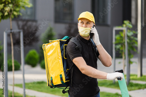 Delivery man in yellow uniform stands with a smartphone against the background of an apartment building, in medical white gloves and facial mask.