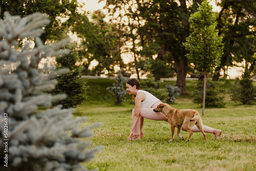 Outdoors yoga at sunrise in public park. Young attractive woman practicing yoga, stretching in Utthan Pristhasana exercise, dog runs nearby.  © Юля Бурмистрова