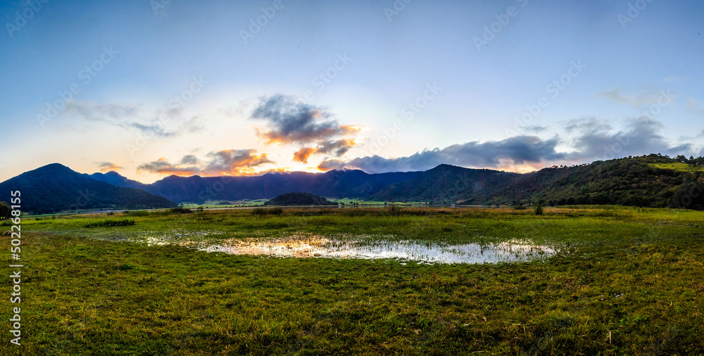 panoramic of volcano crater in the sunset with lake in first plane and mountains in the background, green grass and blue sky in tepetiltic nayarit 