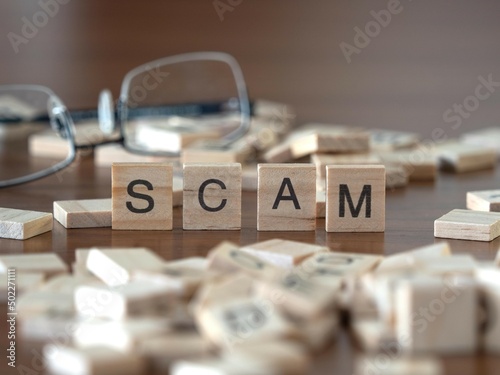 scam word or concept represented by wooden letter tiles on a wooden table with glasses and a book
