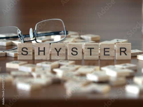 shyster word or concept represented by wooden letter tiles on a wooden table with glasses and a book photo