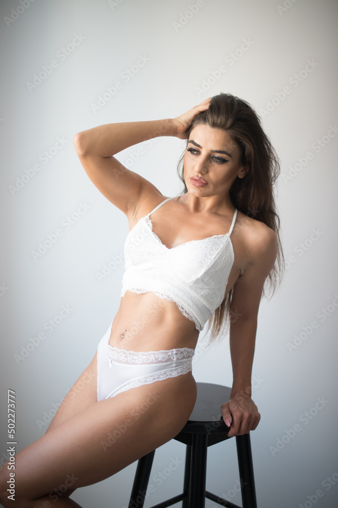 Attractive sexy brunette in white lingerie posing provocatively, in front of a wall ,studio shot. Portrait of sensual woman with long hair, white bikini and bra in classic boudoir scene in studio