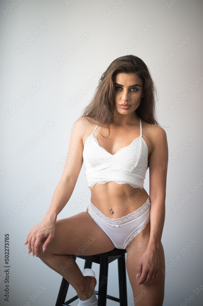 Attractive sexy brunette in white lingerie posing provocatively, in front of a wall ,studio shot. Portrait of sensual woman with long hair, white bikini and bra in classic boudoir scene in studio