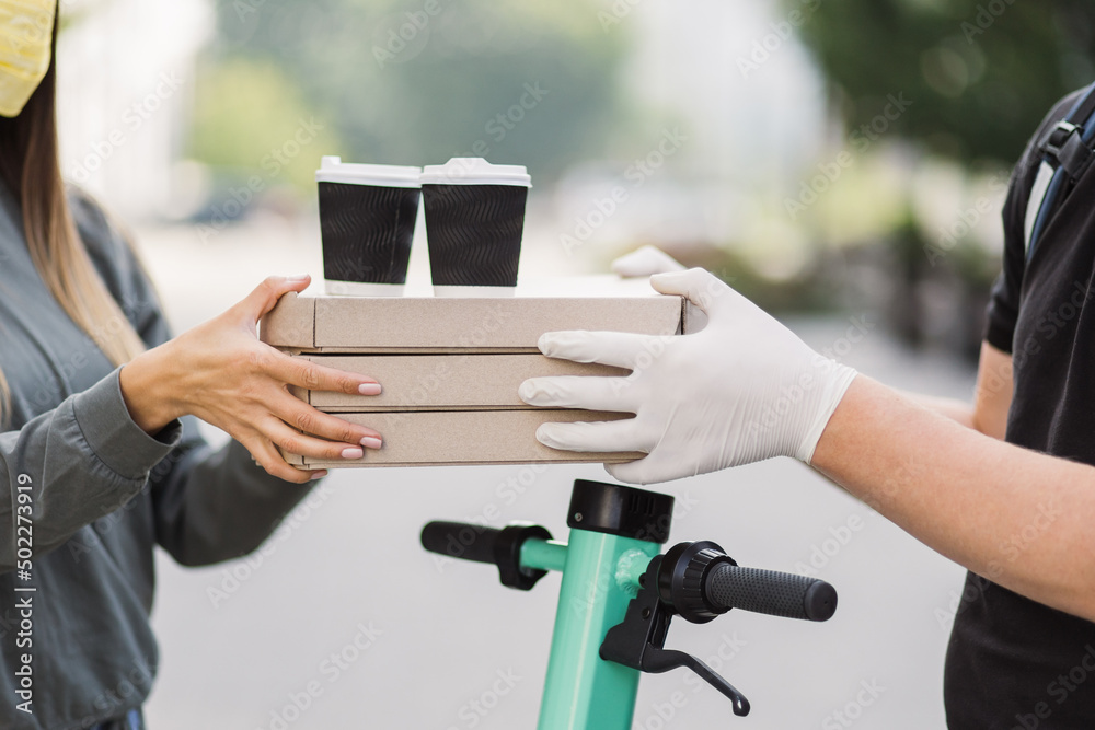 Close up of hands of young man courier in gloves delivering pizza boxes and two cups of coffee to caucasian girl customer in face mask. Delivery man holding cardboard boxes and cups with hot drinks