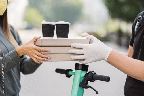 Close up of hands of young man courier in gloves delivering pizza boxes and two cups of coffee to caucasian girl customer in face mask. Delivery man holding cardboard boxes and cups with hot drinks