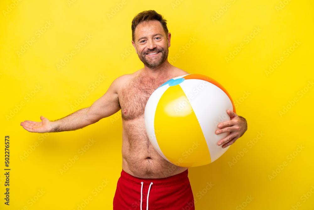 Middle age caucasian man holding beach ball isolated on yellow background extending hands to the side for inviting to come