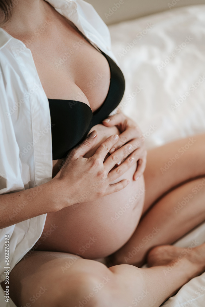 Pregnant woman sits on bed in black bra holding hands on belly Pregnant woman sits on bed in black bra holding hands on belly