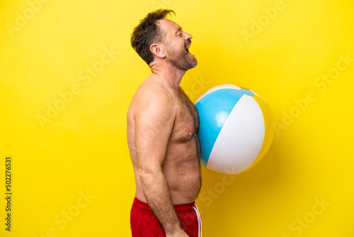 Middle age caucasian man holding beach ball isolated on yellow background laughing in lateral position