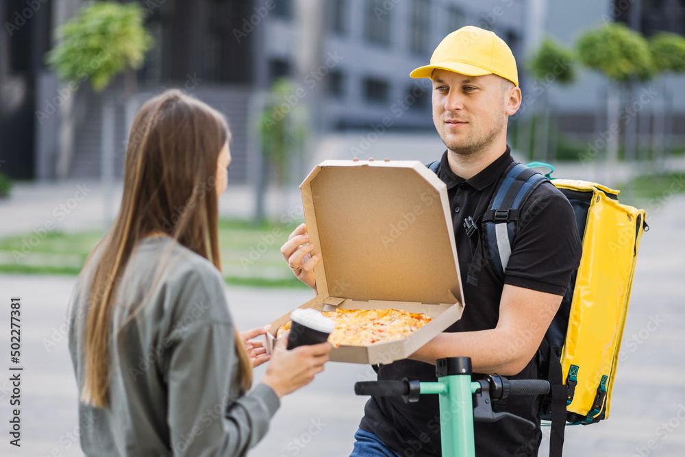 Front view of a delivery man coming to the building of customer and meet young caucasian woman. Girl taking pizza box and cup of coffee from courier. Shipping or delivery service concept.