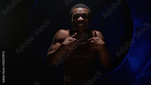 Cool African American teenager with bare, pumped-up body sings hip-hop song and emotionally gestures with his hands. Man raps in music video and sings rap song against blue underground background. photo