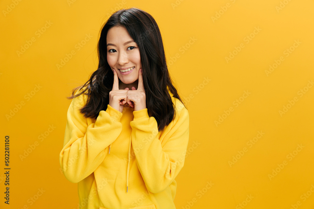 Korean beauty. Cheerful cute Asian student young woman in yellow hoodie sweatshirt touches cheeks by two fingers posing isolated on over orange studio background. Cool offer. People Emotions concept Korean beauty. Cheerful cute Asian student young woman in yellow hoodie sweatshirt touches cheeks by two fingers posing isolated on over orange studio background. Cool offer. People Emotions concept
