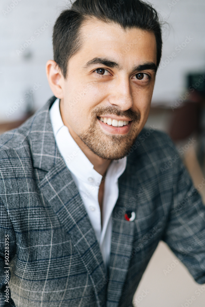 Close-up cropped vertical shot of cheerful elegant business man in fashion suit sitting at desk in modern office room, looking at camera. Front view of serious bearded businessman posing at workplace. Close-up cropped vertical shot of cheerful elegant business man in fashion suit sitting at desk in modern office room, looking at camera. Front view of serious bearded businessman posing at workplace.