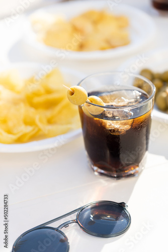 Table full of snacks and a glass of vermuth during a sunny day. Chips, olives and vermut outside with sunglasses on a table. photo