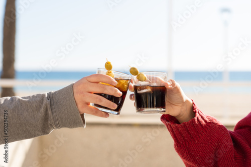 Two hands toasting with their glasses of vermouth in front of the beach during a sunny day. Two people with no faces, clinking with some drinks.