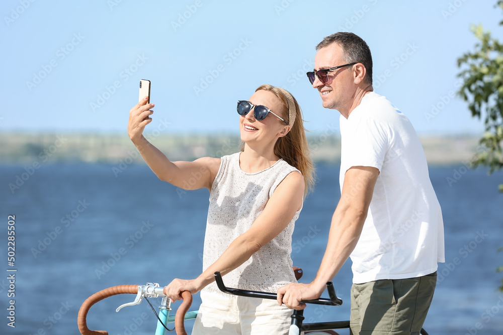 Mature couple with bicycles taking selfie near river on summer day