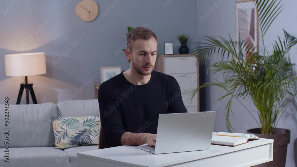 Shot of concentrated young man working on laptop in living room. Guy with beard touching aching neck, sitting at couch. Healthcare concept. Remote work, freelancer