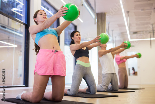 Sports people who are engaged in fitness in the studio for a group workout, perform an exercise on the mat, kneeling and ..holding a mini Pilates ball in front of them