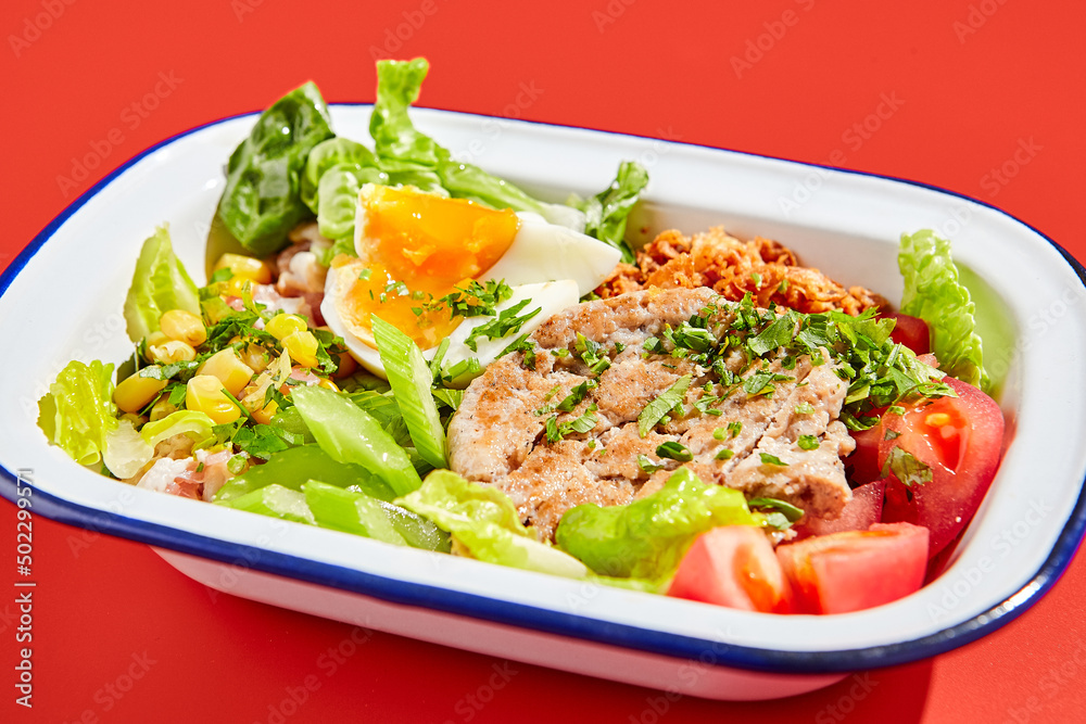 American cobb salad with chicken, eggs and vegetables in minimal style. Healthy salad on trend red background with hard shadow. Bowl with salad, chicken, eggs, vegetables in contemporary style American cobb salad with chicken, eggs and vegetables in minimal style. Healthy salad on trend red background with hard shadow. Bowl with salad, chicken, eggs, vegetables in contemporary style