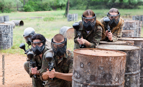 Group of paintball players in masks aiming and shooting with guns at opposing team outdoors