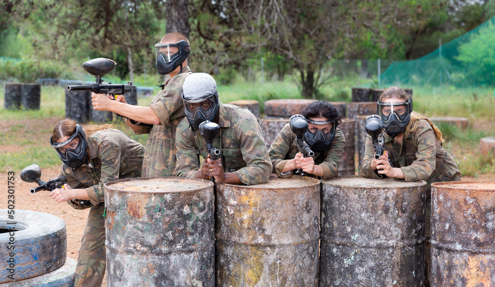 Group of paintball players in masks aiming and shooting with guns at opposing team outdoors