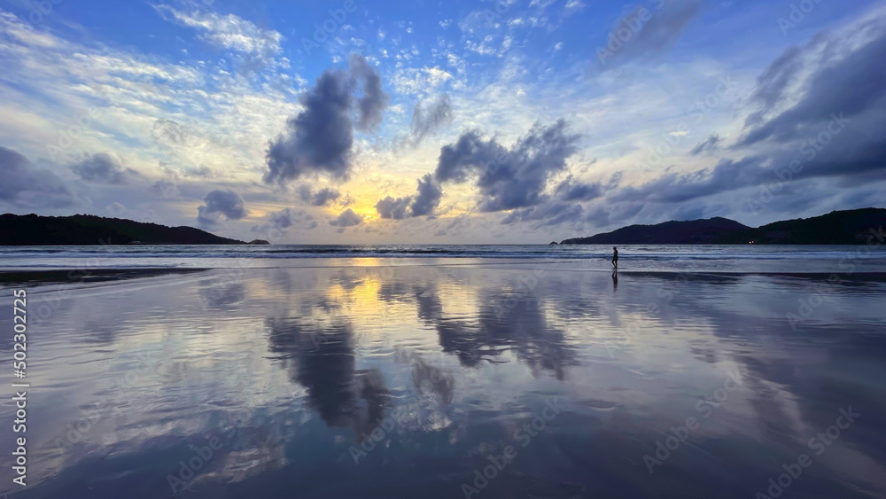 Panorama, seascape, amazing sunset. Twilight, sky with awesome colorful pattern of clouds, reflection on the smooth surface of water. Sunset over the ocean. Man walks alone sea coast. Tropical beach. 