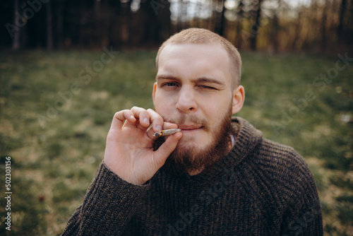 Young man beard smokes cigarette or tobacco. Guy is addicted to cigarettes and always smokes lot of tobacco every day. He smokes cigarette outdoors on background of lake in park. Cool guy blows smoke