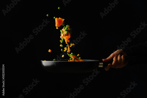 Seafood with vegetables in a frying pan in the hand of a professional chef. Food in a frozen flight on a black background. Sea Asian cuisine. Recipes for restaurant and home cooking.