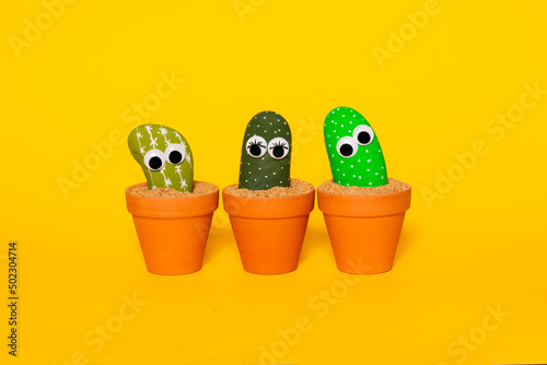 Three painted rocks in shades of green with plastic googly eyes that give a playful and whimsical personification of cactus plants in terra cotta pots on a seamless yellow background